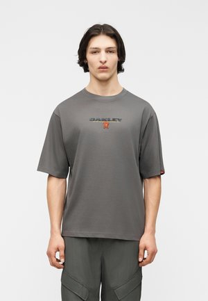 Νέος άνδρας με μεσαίου μήκους σκούρα μαλλιά φοράει μια φαρδιά γκρι μπλούζα Oakley και σκούρο παντελόνι με τσέπες, στέκεται μπροστά σε ένα απλό φωτεινό φόντο.