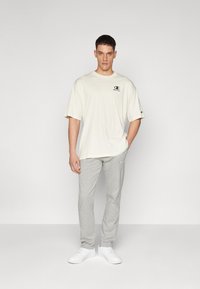 Helles beiges Baumwoll-T-Shirt mit einem kleinen schwarzen Logo auf der Brust, kombiniert mit grauen Jogginghosen und weißen Sneakers. Schlichte, lockere Passform.