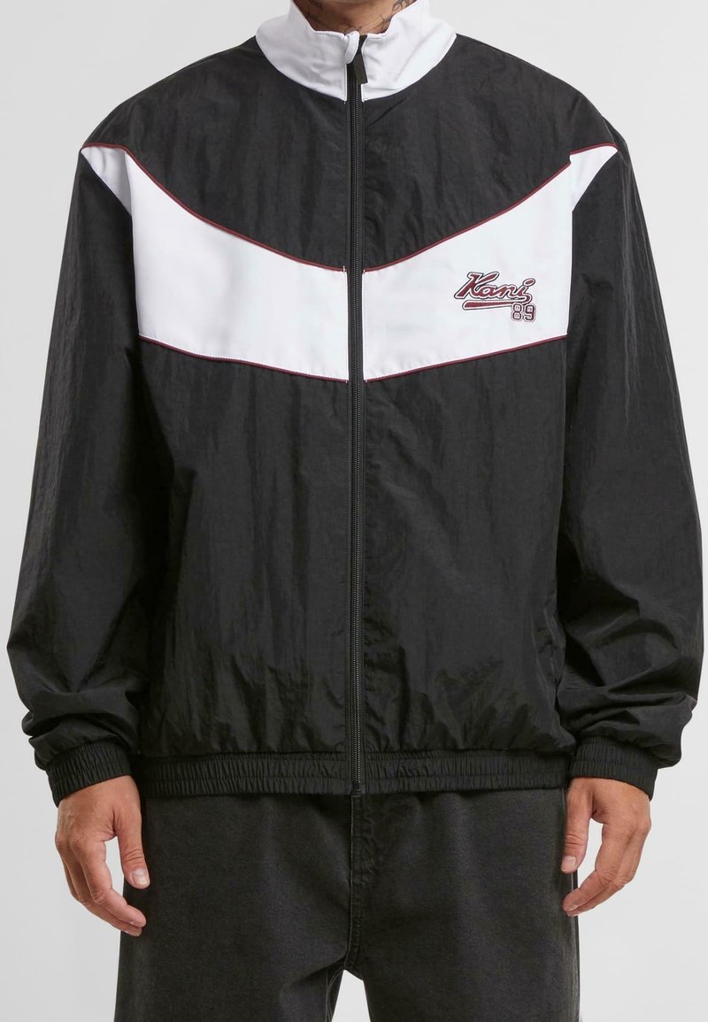 Personne portant une veste coupe-vent zippée noire et blanche avec le logo "Kani 89" sur la poitrine et des poignets et un ourlet élastiques.