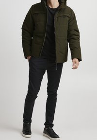 Ciemnozielona kurtka puchowa z kapturem, z przodem na zamek błyskawiczny i podwójnymi kieszeniami na piersi. Nosi ją osoba w czarnej koszuli i czarnych spodniach oraz sneakersach.