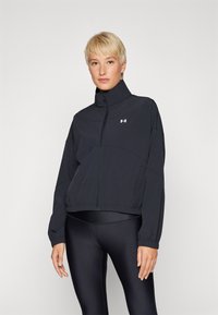 Chaqueta deportiva negra con collar alto, cremallera frontal y mangas largas. Fabricada con un material liviano. Presenta un logo de Under Armour en el pecho.