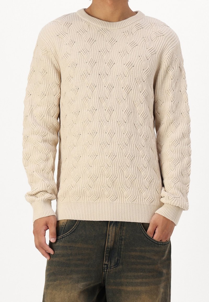 Pull beige en tricot avec un motif torsadé texturé, col rond, poignets et ourlet côtelés. Porté avec un pantalon en denim foncé.