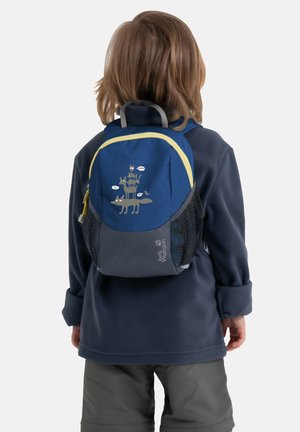 Enfant portant une veste bleu marine et un pantalon vert, portant un sac à dos bleu avec des animaux de dessin animé empilés et des bulles de dialogue à l'arrière.