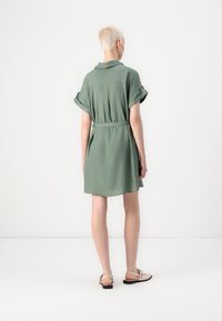 Abito verde a lunghezza ginocchio con maniche arrotolate, colletto e vita con cintura. Realizzato in tessuto morbido, con una texture liscia e un design semplice.