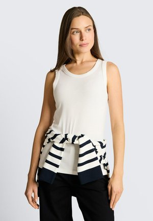 Kobieta w białym, bezrękawnym topie i czarnych spodniach z paskowanym granatowo-białym swetrem związanym w talii, patrząca w bok.