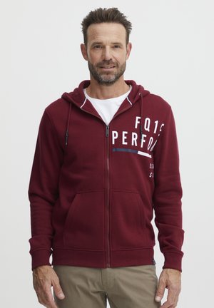 Burgunderfarget hoodie med glidelås, snøring og frontlommer. Har hvit trykt tekst og en myk, teksturert overflate.