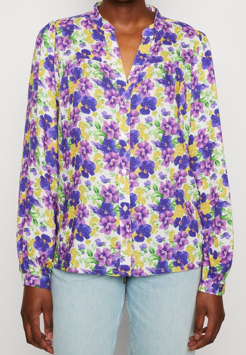 Blouse florale avec un décolleté en V, ornée de pensées violettes et d'accents jaunes sur un fond blanc. Manches longues avec poignets à boutons.