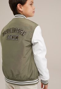 Oliwkowa bomberka z białymi rękawami, żebrowanym kołnierzem i pasem w talii. Na plecach znajduje się wyszyty napis "BLUE RIDGE DENIM".