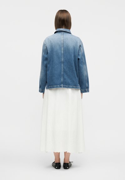 Γυναίκα που κοιτάζει προς τα πίσω, φοράει μπλε τζιν μπουφάν πάνω από άσπρο μίντι φόρεμα και μαύρα επίπεδα παπούτσια με ασημένια καρφιά.