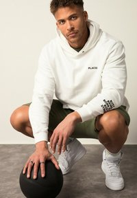 Sudadera blanca con "PLACID" en negro en el pecho y texto en la manga. Pantalones cortos verde oliva, zapatillas blancas y balón de baloncesto negro sobre una superficie gris.