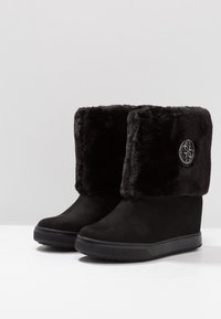 Botas de gamuza negra con cuñas y gruesos puños de pelaje sintético negro, así como un logotipo metálico redondo en el lado exterior de cada bota.