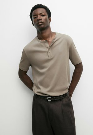 Man met kort krullend haar, gekleed in een beige Henley met korte mouwen en een donkere broek met een zwarte riem, staand tegen een witte muur.