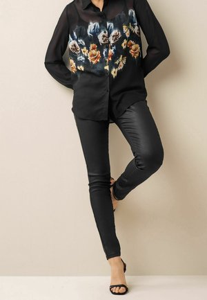 Chemise noire à imprimé floral sur le devant, associée à un pantalon noir ajusté. La chemise est dotée de boutons et de manches longues. Sandales noires à bouts ouverts.