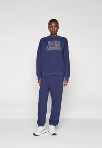 Mørkeblå sweatpants og sweatshirt med "UNDER ARMOUR" logo i hvid på brystet, med en afslappet pasform og ribbestrikkede manchetter.