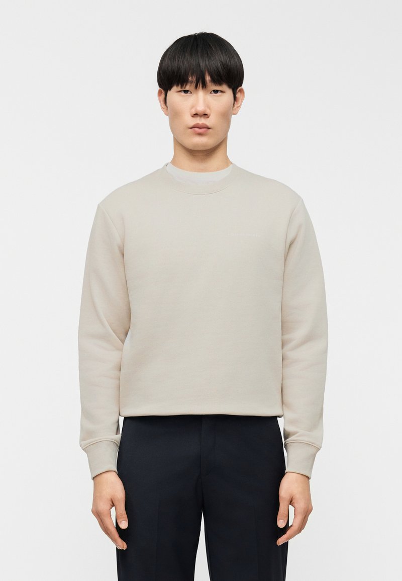 Φούτερ σε ανοιχτό μπεζ χρώμα με μακριά μανίκια και στρογγυλή λαιμόκοψη, κατασκευασμένο από απαλό ύφασμα. Διαθέτει διακριτικό λογότυπο στο μπροστινό μέρος και ριμπ τελειώματα στα μανίκια.