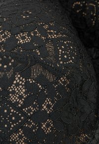 Svart spets tyg med blommönster och öppna vävningar, med en strukturerad yta och kontrasterande beige bakgrund.