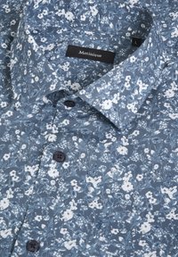 Chemise bleue à boutons avec motif floral blanc et boutons noirs, étiquette Matinique, taille large, présentée pliée avec col visible.