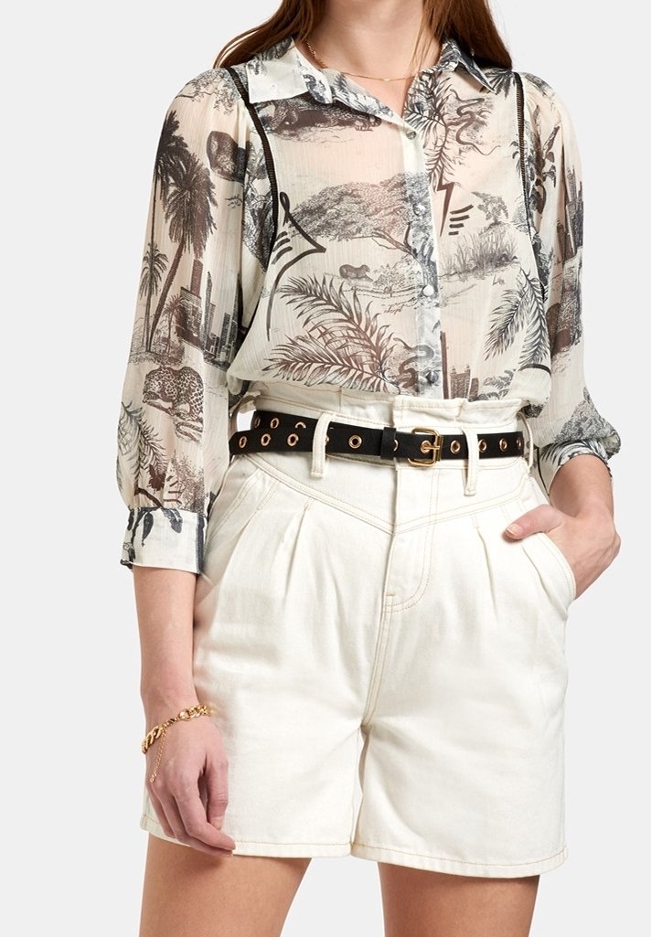 Frau trägt ein durchsichtiges, gemustertes Hemd mit Tier- und Stadtmotiven, kombiniert mit hoch taillierten weißen Shorts und einem schwarzen Gürtel.