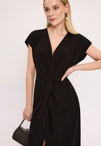Czarna sukienka typu wrap z krótkimi rękawami, z głębokim dekoltem w kształcie V, związaną w talii oraz z rozcięciem z boku. Nosi ją z koralikową torebką.