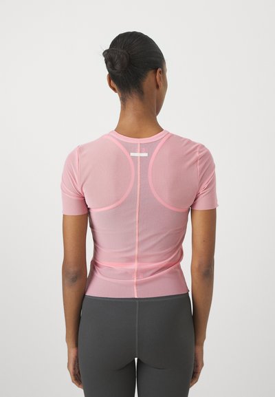 Rövid ujjú, testhezálló sportfelső világos rózsaszín hálós anyagból, sima textúrával és diszkrét hátsó varrással. Sötétszürke leggings-szel párosítva.
