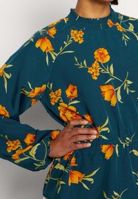 Blommig blus i teal med gula-orange blommor, samlad halsringning och ärmslut, elastisk midja, slät tyg och långa ärmar.