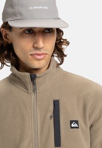 Beige fleece jacket with a high collar, zippered front, and black accents. Grey cap with logo and a flat brim. Textured fabric.