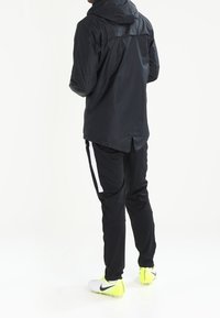 Black sports jacket with hood, paired with black pants featuring white side stripes and bright yellow soccer cleats. Smooth textile finish.