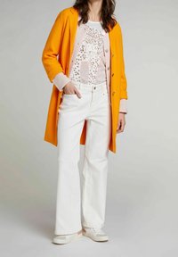 Abrigo de lana naranja sobre una blusa de capas rosa claro, combinado con jeans acampanados blancos y zapatillas beige con detalles en plata.