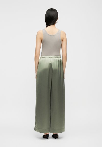 Γυναίκα που στέκεται με την πλάτη γυρισμένη, φοράει γκρι αμάνικη κορυφή με ριμπ ύφανση, ανοιχτό πράσινο σατέν παντελόνι με φαρδιά μπατζάκια και μαύρα ψηλά τακούνια.