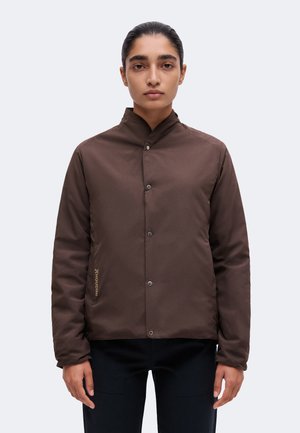 Personne aux cheveux foncés attachés porte une veste marron boutonnée et un pantalon noir, debout devant un fond clair uni.