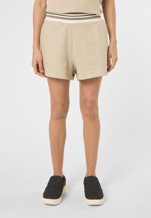 Frau trägt beige Shorts und passendes Oberteil mit gestreiftem Taillenbund, kombiniert mit schwarzen Slip-On-Schuhen mit weißen Sohlen.