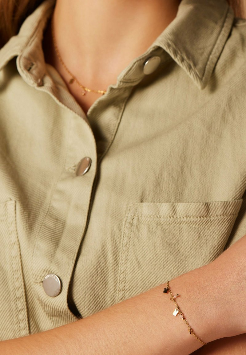 Gouden armband met kleine geometrische bedels, gedragen op een arm met een lichte huidskleur. De achtergrond toont een lichtgroene gestreepte blouse.