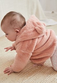 Chandail à capuche en peluche rose doux avec une capuche arrondie et de petites oreilles, porté par un bébé qui rampe sur une surface texturée de couleur claire.