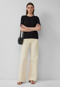 Schwarzes, tailliertes T-Shirt kombiniert mit cremefarbenen, weiten Hosen. Das Model hält eine kleine, strukturierte schwarze Handtasche und steht vor einem neutralen Hintergrund.