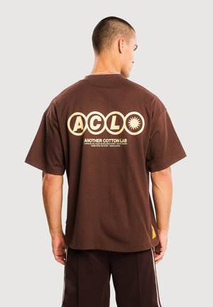 Giovane uomo con capelli corti che indossa una t-shirt marrone oversize con "ACLO" e "Another Cotton Lab" stampati in beige sul retro.
