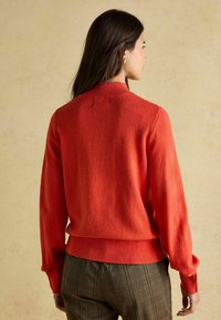 Roter Strickpullover mit hohem Kragen, gerippten Bündchen und Saum. Die Textur wirkt weich und wird über karierter Hose getragen.