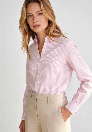 Vrouw met lichtbruin haar die een roze-wit gestreept overhemd draagt, ingestopt in beige hoogtalige broek met haar hand in de zak.