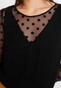 Černý top s průhledným síťovaným výstřihem s černými puntíky a texturovaným, jednobarevným černým tělem. Design s véčkovým výstřihem a dlouhými rukávy.