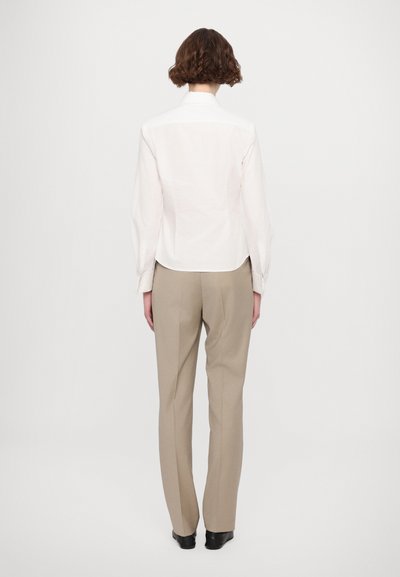 Camicia bianca in cotone con colletto, polsini abbottonati e orlo curvo, abbinata a pantaloni beige su misura e scarpe nere, vista dal retro.