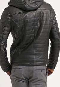 Giacca di pelle nera con cappuccio, caratterizzata da un design trapuntato, una forma aderente e dettagli di cucitura visibili sul retro. Abbinata a pantaloni grigi.