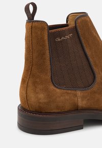 Bottine en daim marron avec un panneau élastique en chevrons, des garnitures en cuir et une semelle en bois foncé contrastante. Dotée d'une languette pour un enfilage facile.