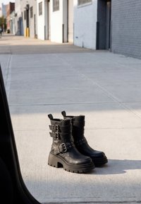 Μαύρα δερμάτινα μποτάκια αστραγάλου με χοντρούς τακούνια και πολλαπλές αγκράφες στέκονται σε έναν φωτεινό αστικό πεζόδρομο κοντά σε ένα κτίριο.