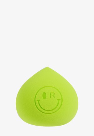 Spugna per trucco verde a forma di goccia con un volto sorridente che strizza l'occhio e le lettere "T" e "R" su sfondo bianco.