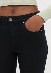 Črne kavbojke iz jeansa s slim krojem, sprednjim žepom, srebrnim gumbom in subtilnimi detajli šivov, s gladko teksturo in srednje visokim pasom.