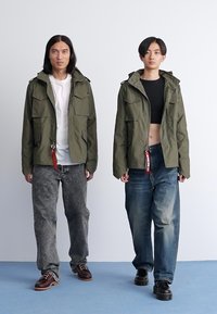 Két modell olajzöld dzsekiket visel, amelyeken több zseb található, laza farmernadrággal párosítva. A bal oldali modellen szürke póló van, a jobb oldalin pedig fekete cropped top.