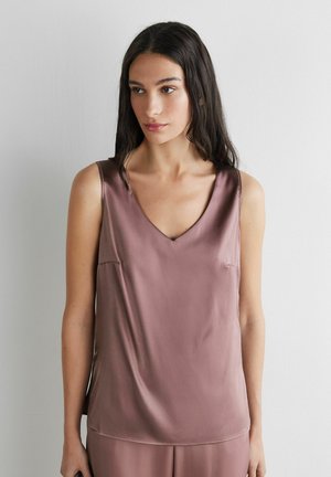 Kvinde med langt mørkt hår iført en ærmeløs mauve satin V-hals top mod en ensfarvet hvid baggrund.