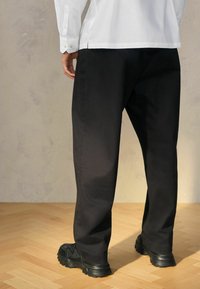 Pantalon en coton noir à coupe décontractée, avec une texture lisse, des jambes droites, assorti à des chaussures noires texturées.