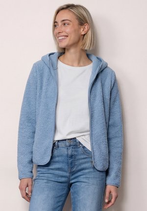 Kvinde med kort blondt hår, smilende, iført en lyseblå fleecejakke over en hvid skjorte og blå jeans mod en ensfarvet baggrund.