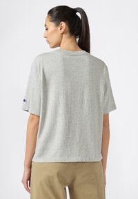 Camiseta de manga corta gris hecha de una suave mezcla de algodón. Presenta un corte relajado y un logo de Champion en la manga izquierda, con un escote redondo.