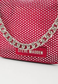 Rode handtas met strassversieringen, zilveren schouderband en een merkteken. Gekartelde oppervlakte met een patroon van kleine glanzende studs.
